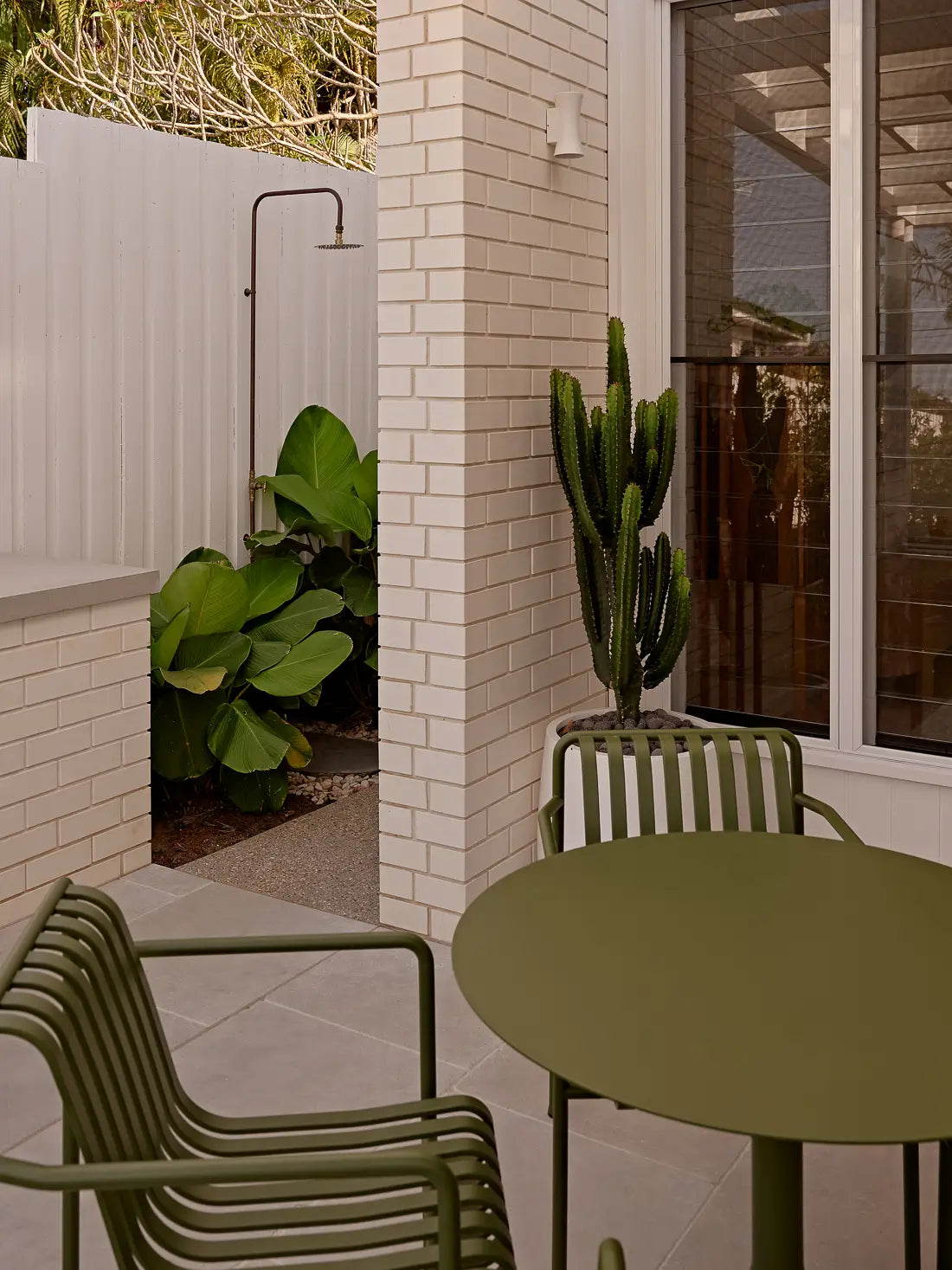 Outdoor shower and patio corner with green chairs, cactus, and white brick wall.