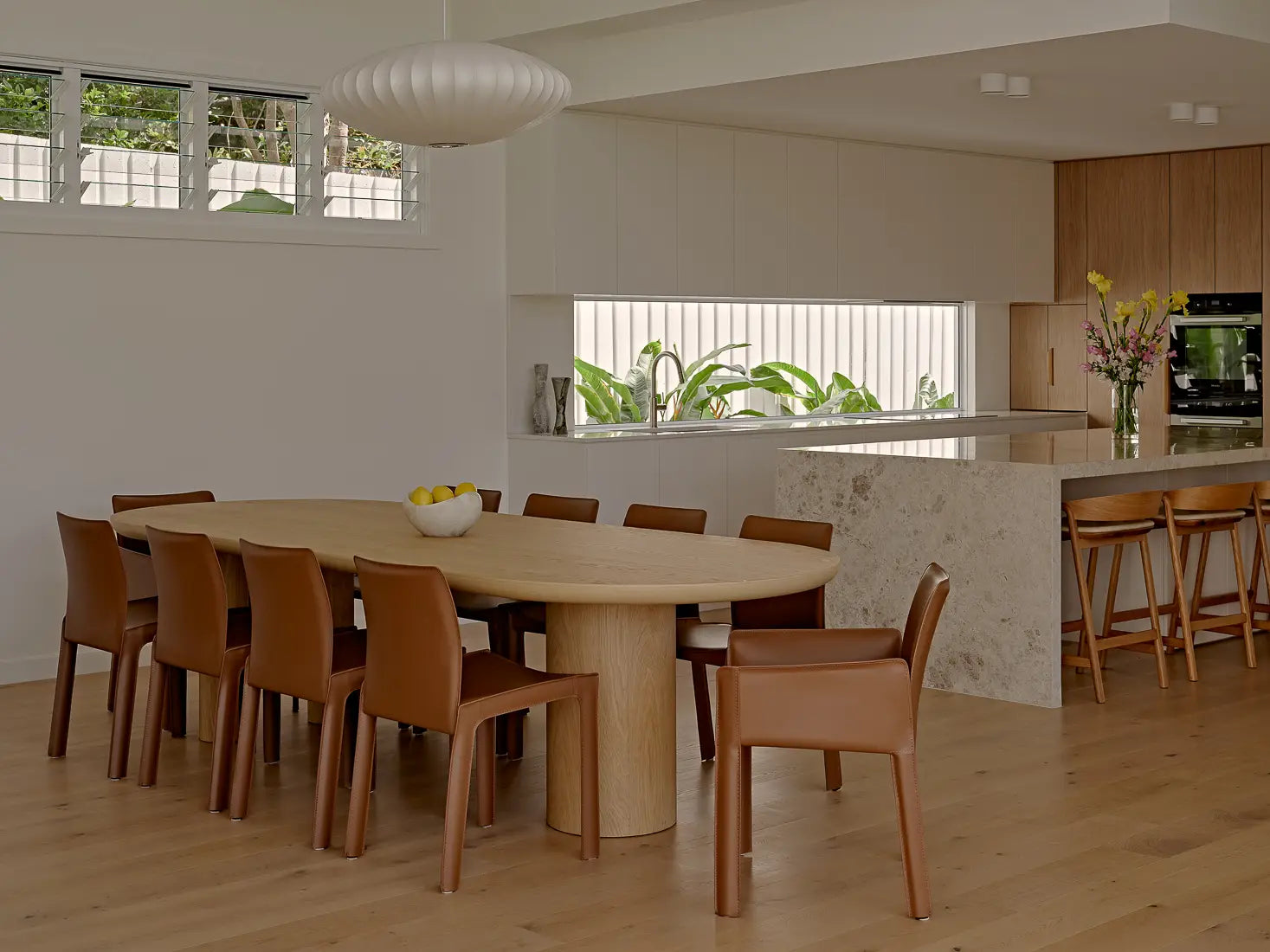 Dining area with a long wooden table, leather chairs, and view into a modern kitchen.