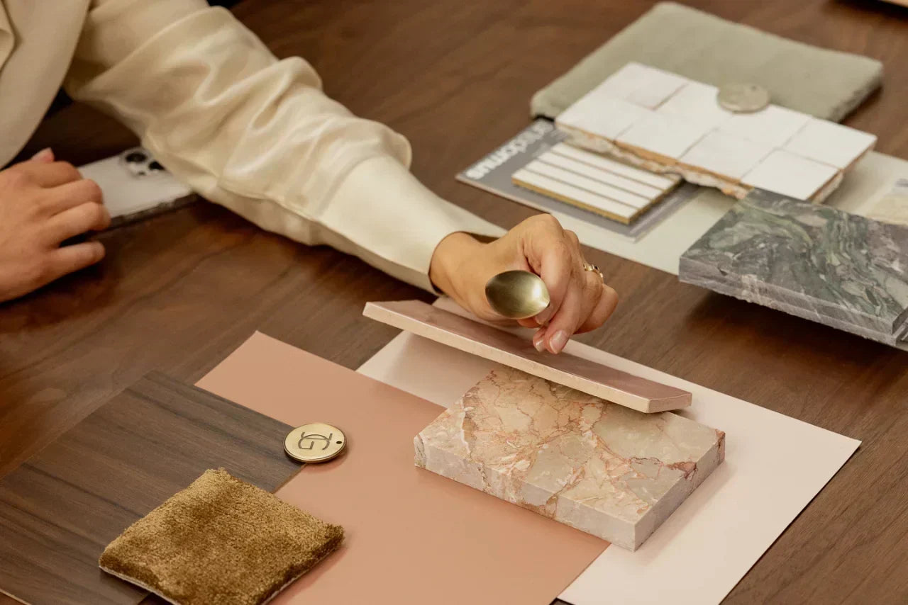 Hands arranging interior design material samples, including marble swatches, tiles, wood finishes, and brass hardware on a mood board