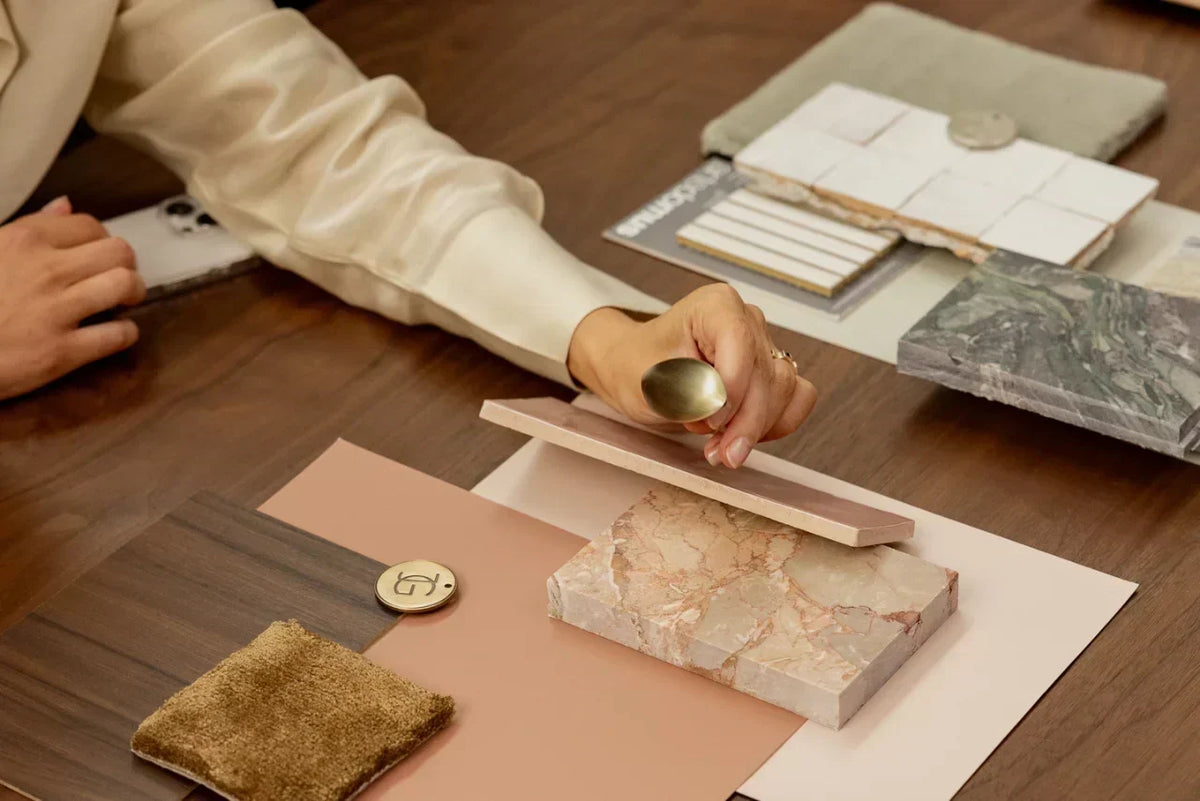 Hands arranging interior design material samples, including marble swatches, tiles, wood finishes, and brass hardware on a mood board