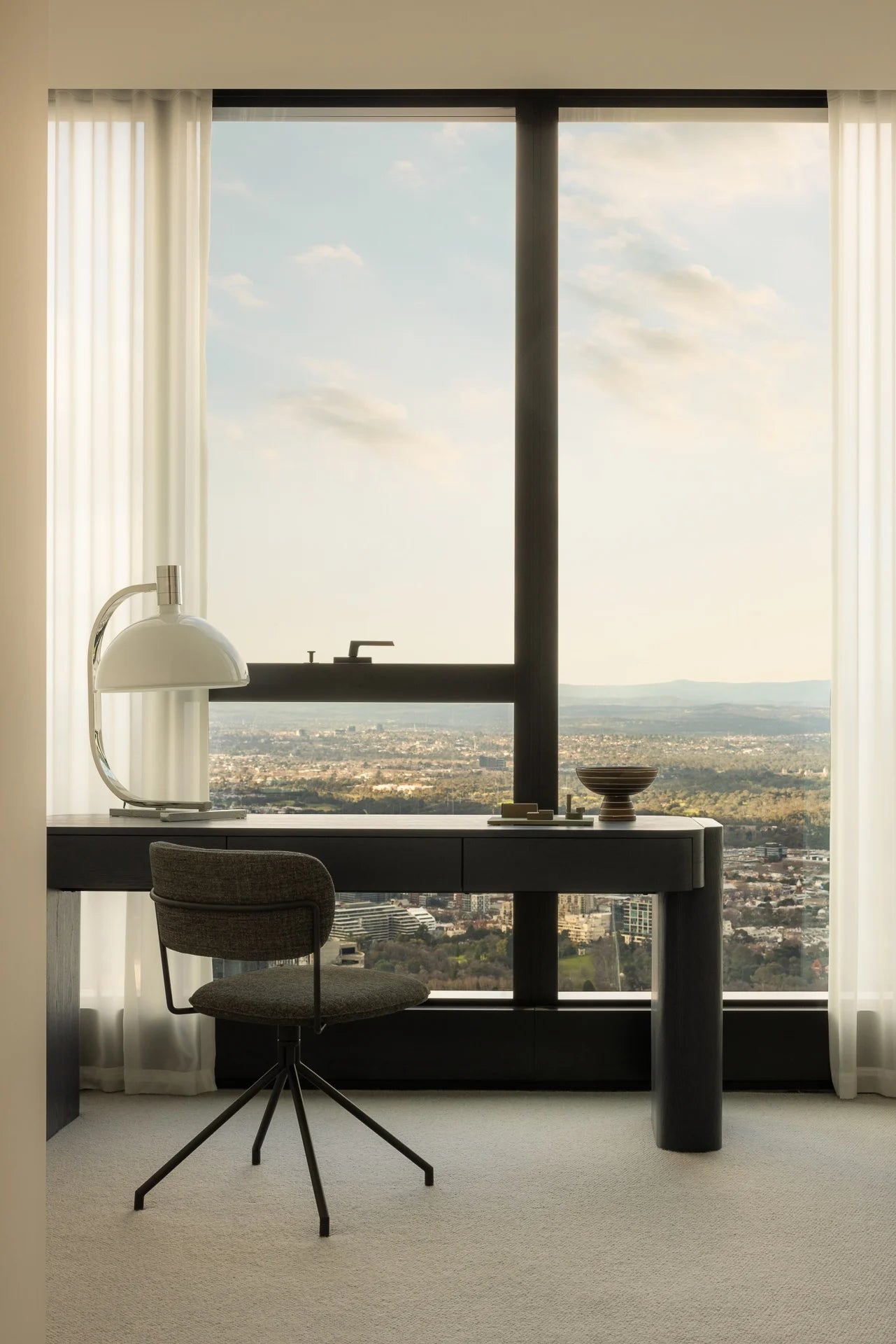 Modern office desk with chair in front of large windows with a cityscape view.